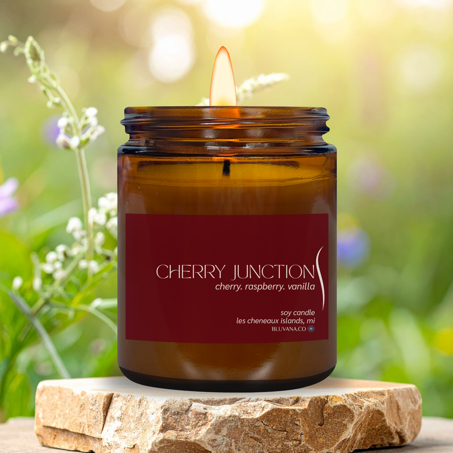 Candle in an amber jar with a red label on a stone surface, surrounded by greenery.