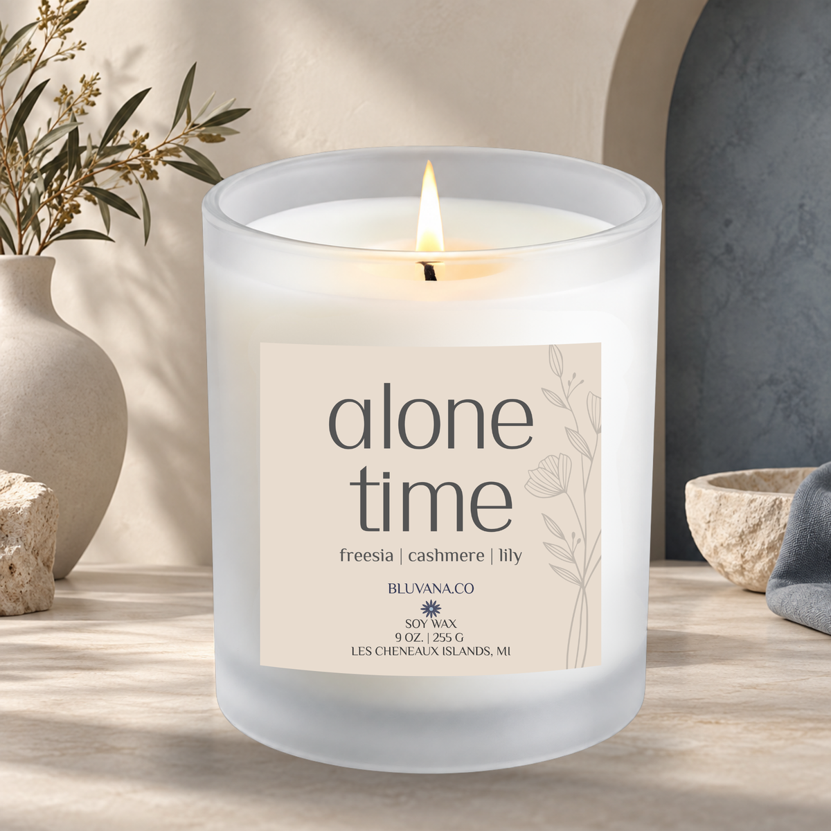 Image of a white frosted glass vessel with flame burning sitting on a modern countertop with ceramic vase in background. Label is a creamy beige that reads Alone Time with scent notes of freesia, cashmere and ily