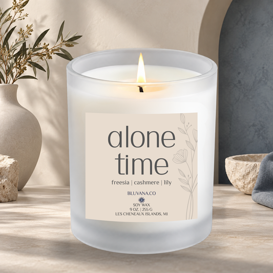 Image of a white frosted glass vessel with flame burning sitting on a modern countertop with ceramic vase in background. Label is a creamy beige that reads Alone Time with scent notes of freesia, cashmere and ily