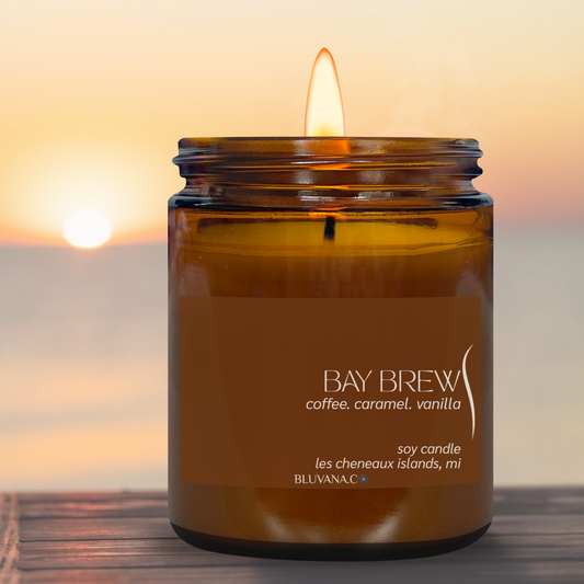 A lit amber glass soy candle jar with a brown label reading 'BAY BREW coffee, caramel, vanilla'', set on a wooden surface against a blurred sunset seascape background.