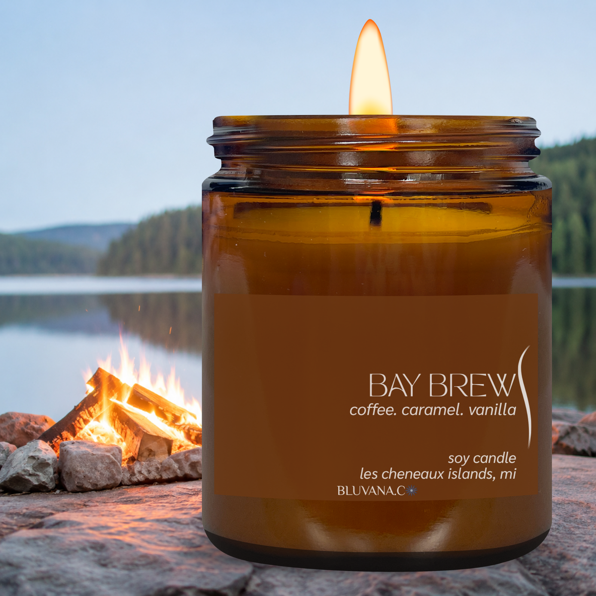 A lit amber glass soy candle jar with a brown label reading 'BAY BREW coffee, caramel, vanilla'', set on a flat rocky surface against a lake and bonfire seascape background.