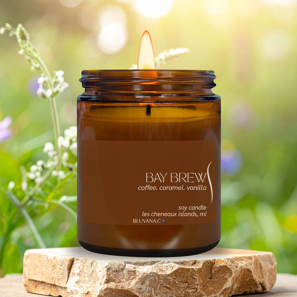 An amber glass jar candle with a lit flame sits on a rustic stone slab outdoors. The label reads 'BAY BREW coffee, caramel, vanilla'.