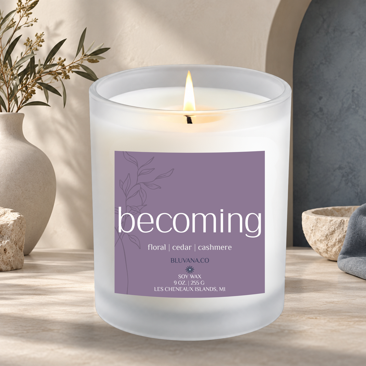 Image of a white frosted glass candle vessel with flame burning sitting on a modern countertop with ceramic vase in background. Label is a muted purple that reads Becoming with scent notes of floral cashmere and cedar.