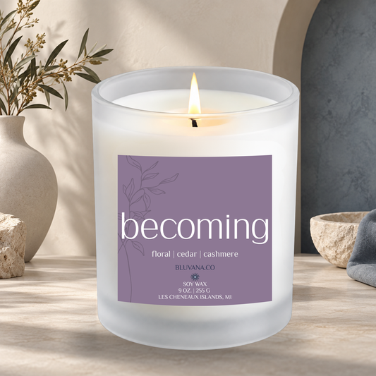 Image of a white frosted glass candle vessel with flame burning sitting on a modern countertop with ceramic vase in background. Label is a muted purple that reads Becoming with scent notes of floral cashmere and cedar.
