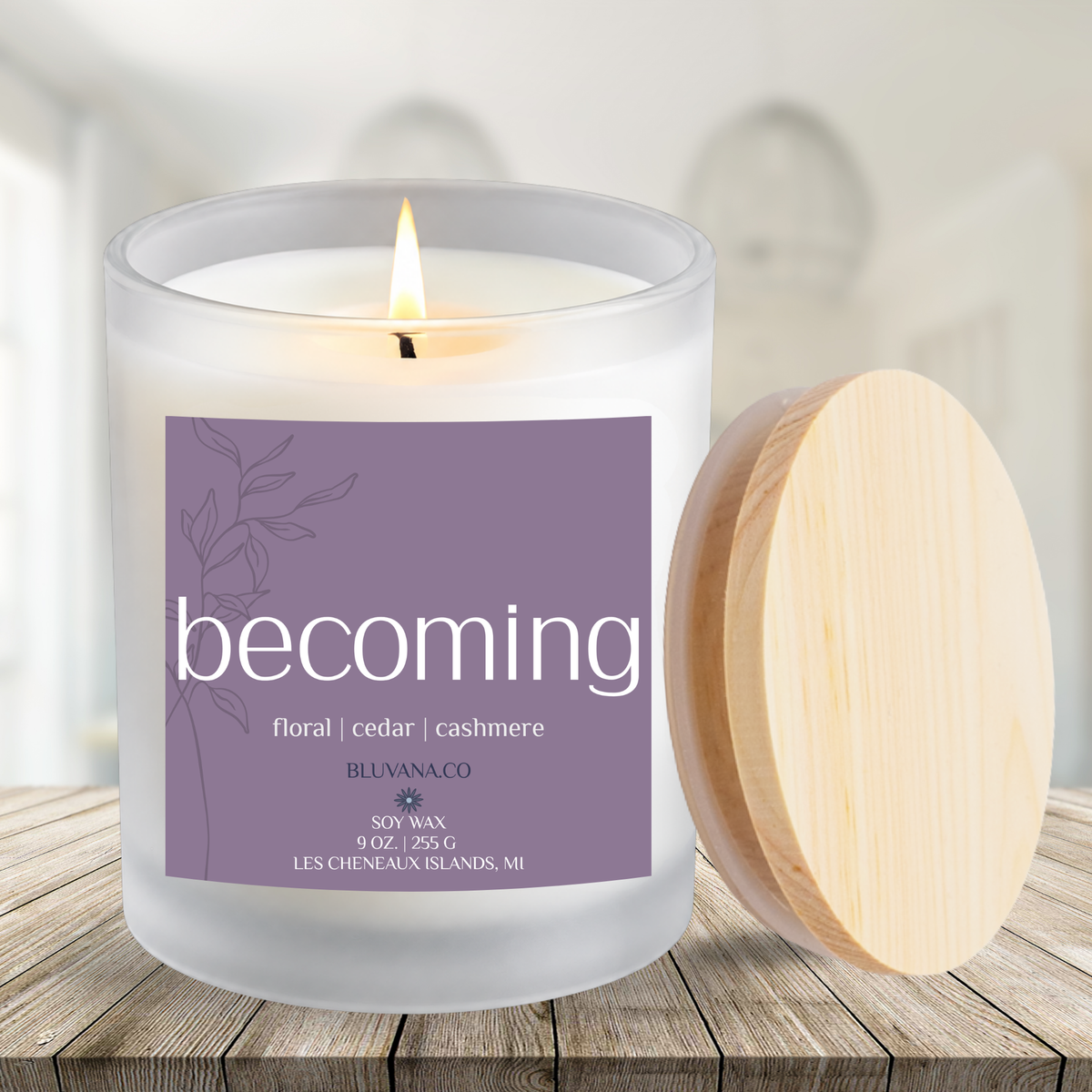 Image of a white frosted glass candle vessel with flame burning sitting on a wood countertop with minimalistic white background. Label is a muted purple that reads Becoming with scent notes of floral cashmere and cedar.