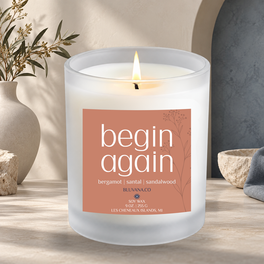 Image of a white frosted glass vessel with flame burning sitting on a modern countertop with ceramic vase in background. Label is rusty orange that reads begin Again with scent notes of bergamot, santal, and sandalwood.