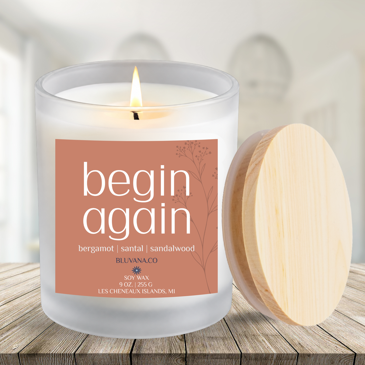 Image of a white frosted glass vessel with flame burning sitting on a modern wood countertop. Label is rusty orange that reads begin Again with scent notes of bergamot, santal, and sandalwood.