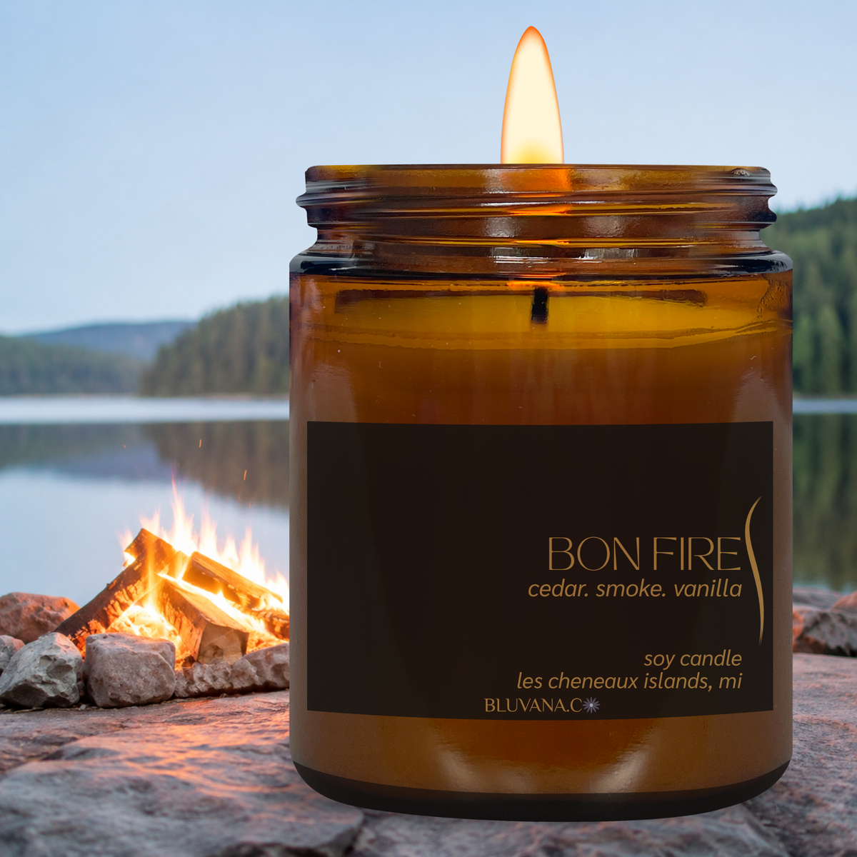 A lit amber glass jar candle labeled BON FIRE, shown in front of a campfire with a lake and forest background, suggesting a outdoor camping scent.