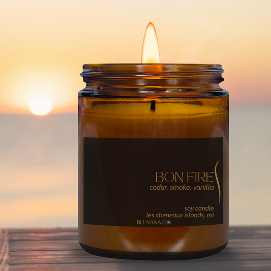 An amber glass jar candle with a lit flame, featuring a black label with gold text that reads 'BON FIRE cedar. smoke.