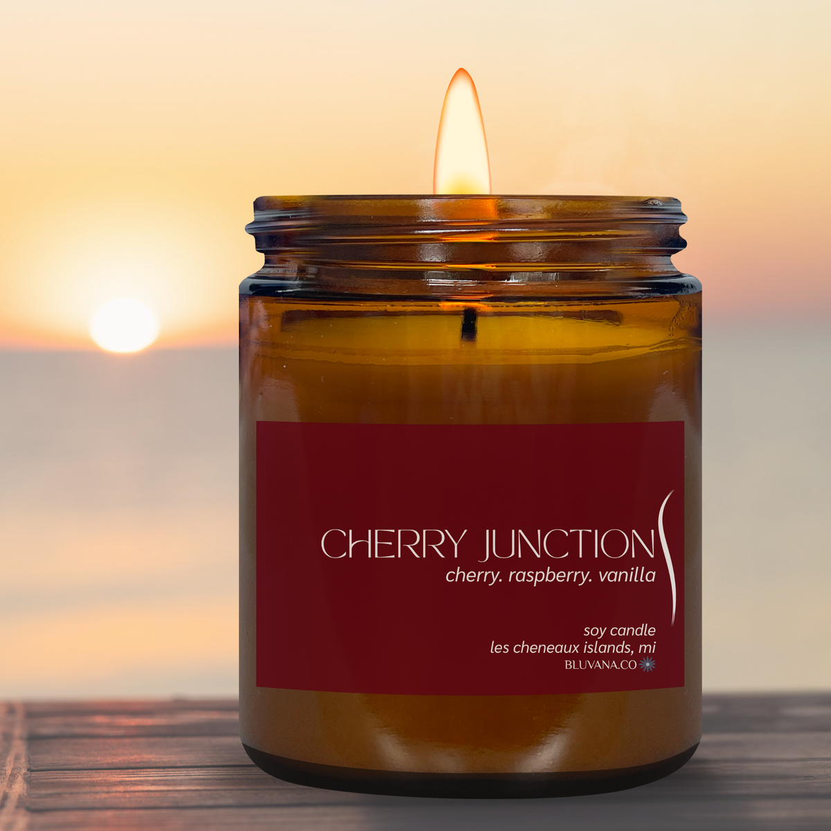 A lit amber glass jar candle sits on a wooden surface with a blurred sunset seascape in the background. The label reads CHERRY JUNCTION with scent text including cherry, raspberry, and vanilla.