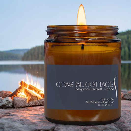 Candle in an amber jar with a label 'Coastal Cottage' by a campfire by a lake.