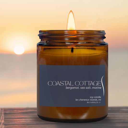 A lit amber glass jar candle sits on a wooden surface with a blurred sunset seascape in the background. The label reads COASTAL COTTAGE with scent text including bergamot, sea salt, and marine.