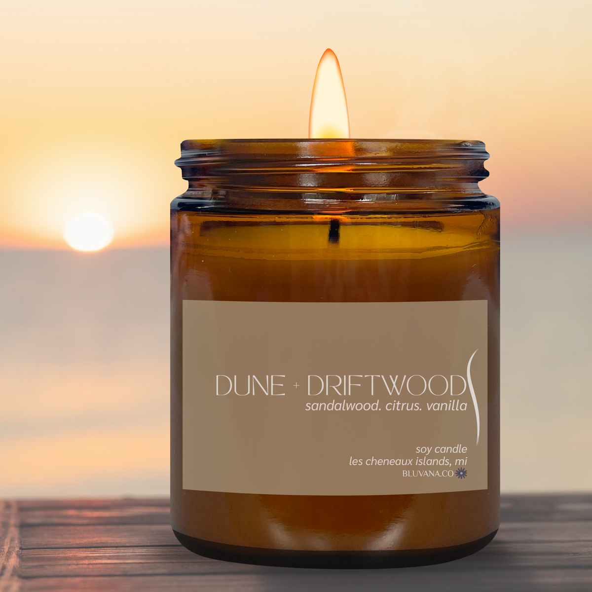 Amber glass candle jar with khaki label that reads DUNE + DRIFTWOOD. Candle is sitting on a dock with a sunset in the background. 