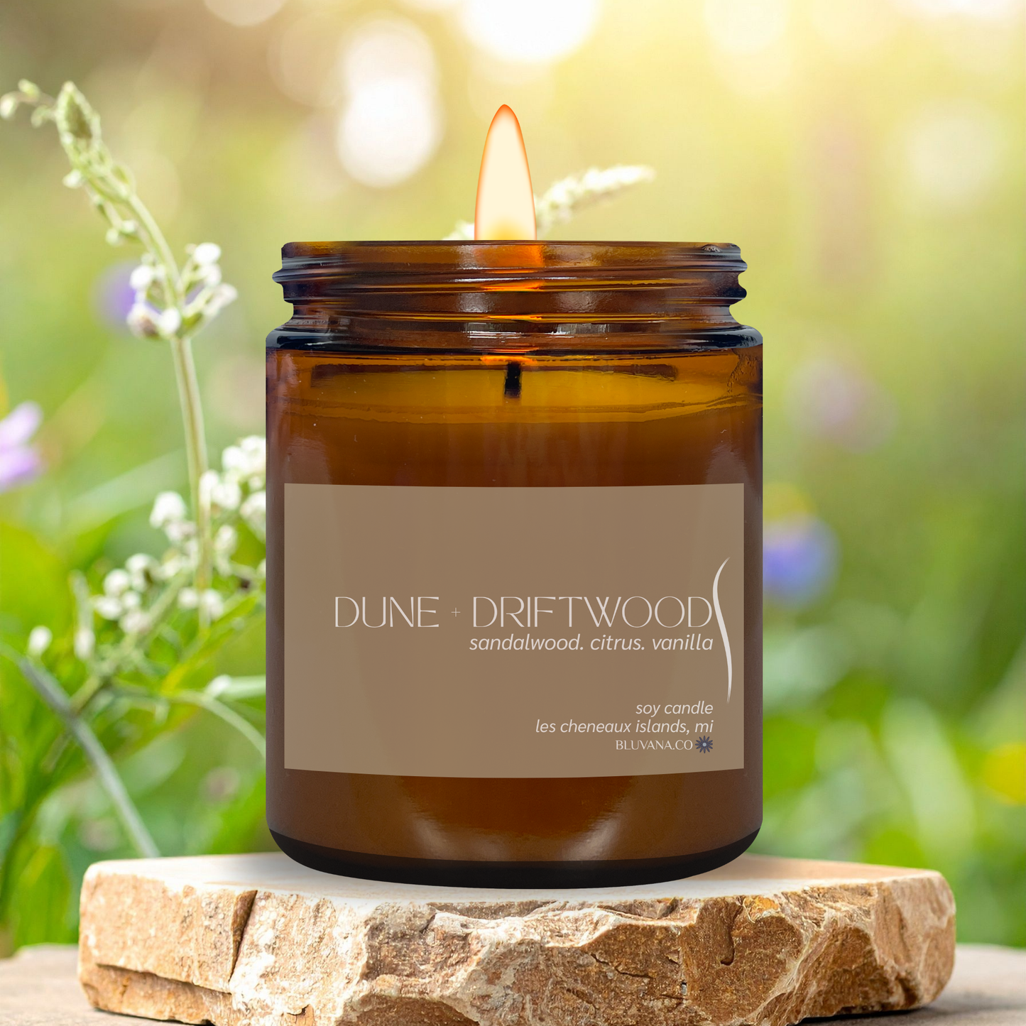 Brown candle in a jar labeled 'Dune Driftwood' on a stone surface with a natural background.