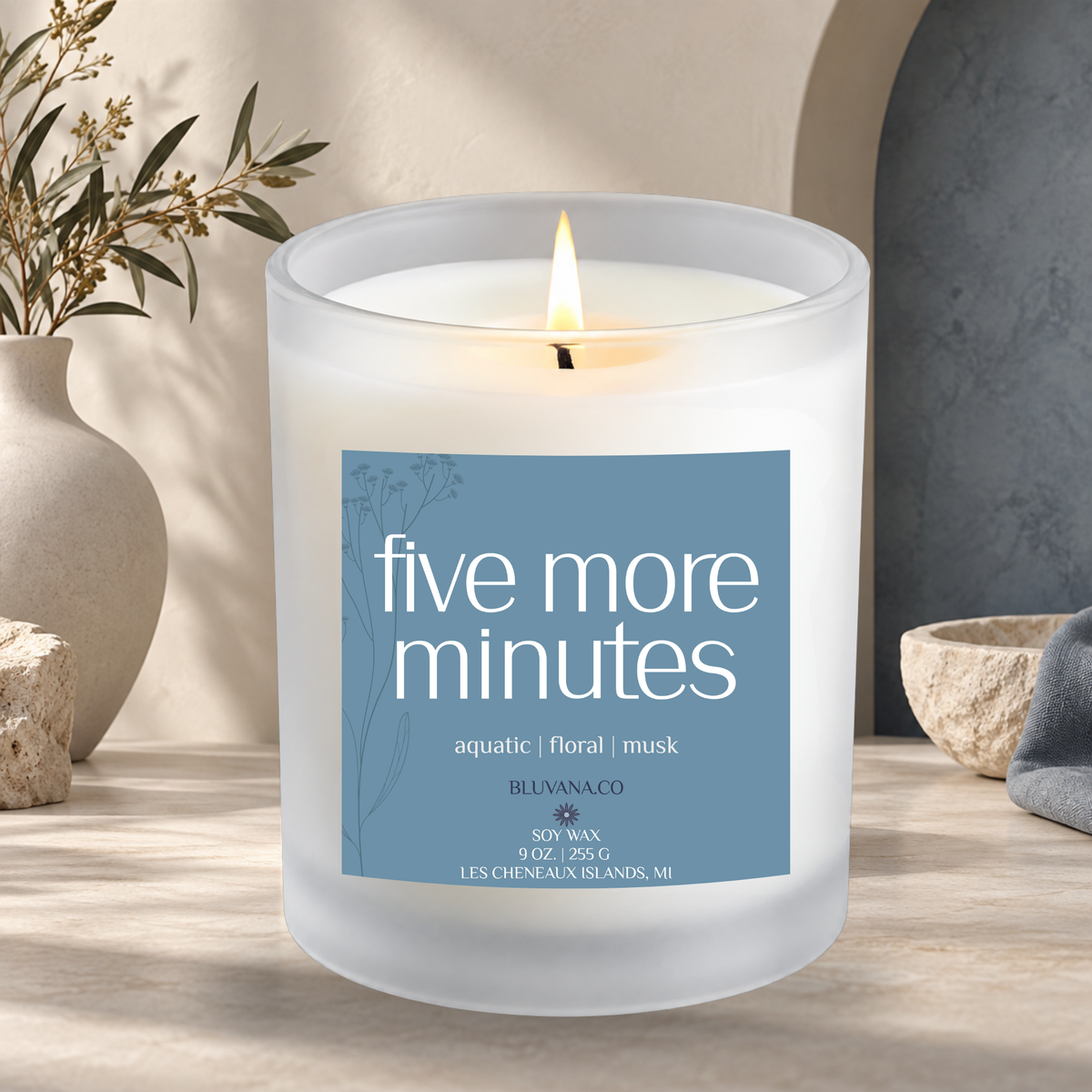 Image of a white frosted glass vessel with flame burning sitting on a modern countertop with ceramic vase in background. Label is a blue gray color that reads Five More Minutes with scent notes of aquatic, floral, and musk. 
