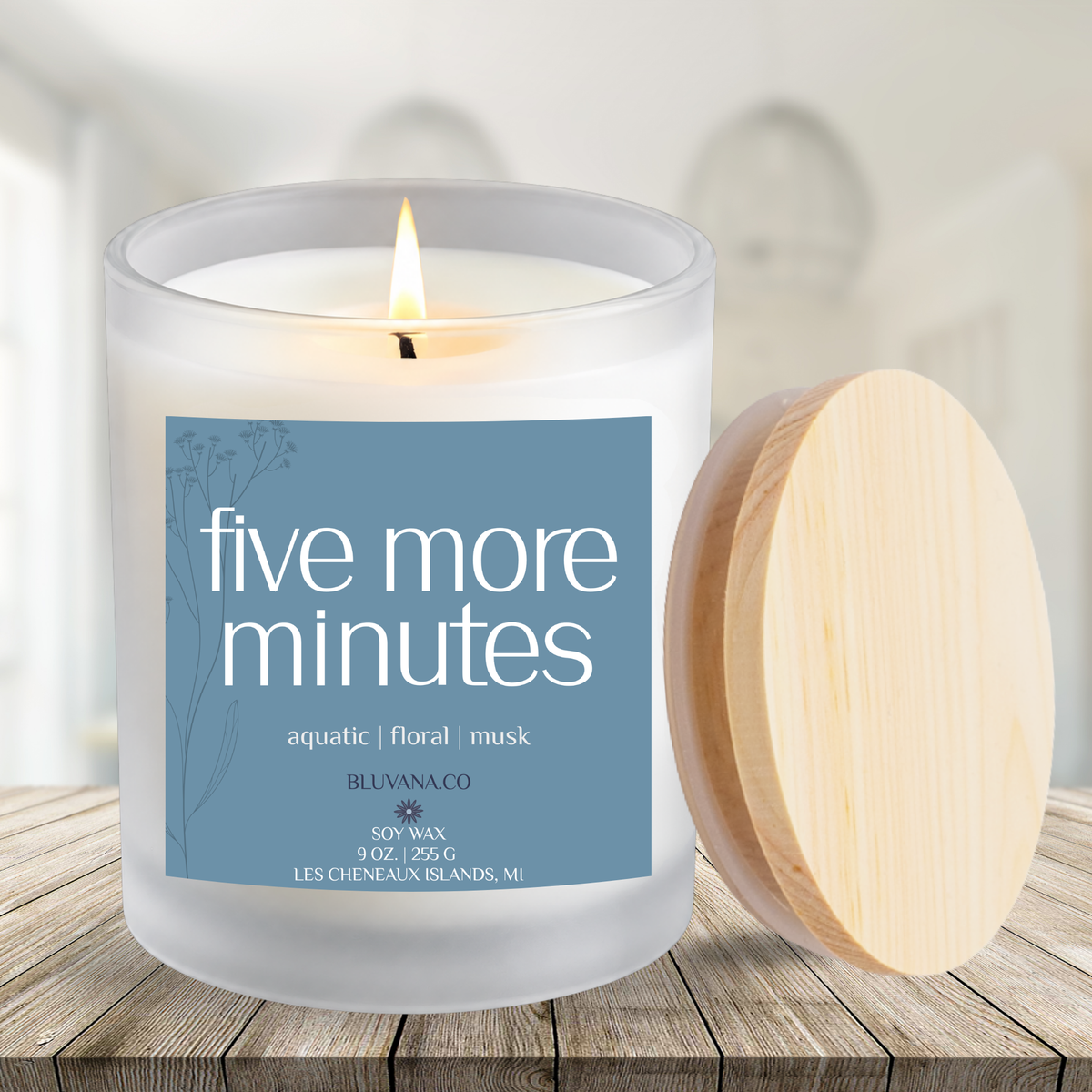 Image of a white frosted glass vessel with flame burning sitting on a modern wood countertop. Label is a blue gray color that reads Five More Minutes with scent notes of aquatic, floral, and musk. 
