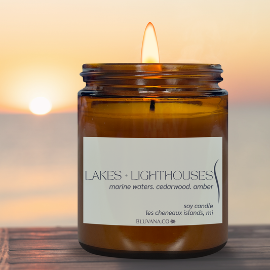 amber jar candle with light blue label that reads Lakes + Lighthouses, featuring a aquatic water, cedarwood fragrance. Jar is situated on a tale with sunset in the background. 