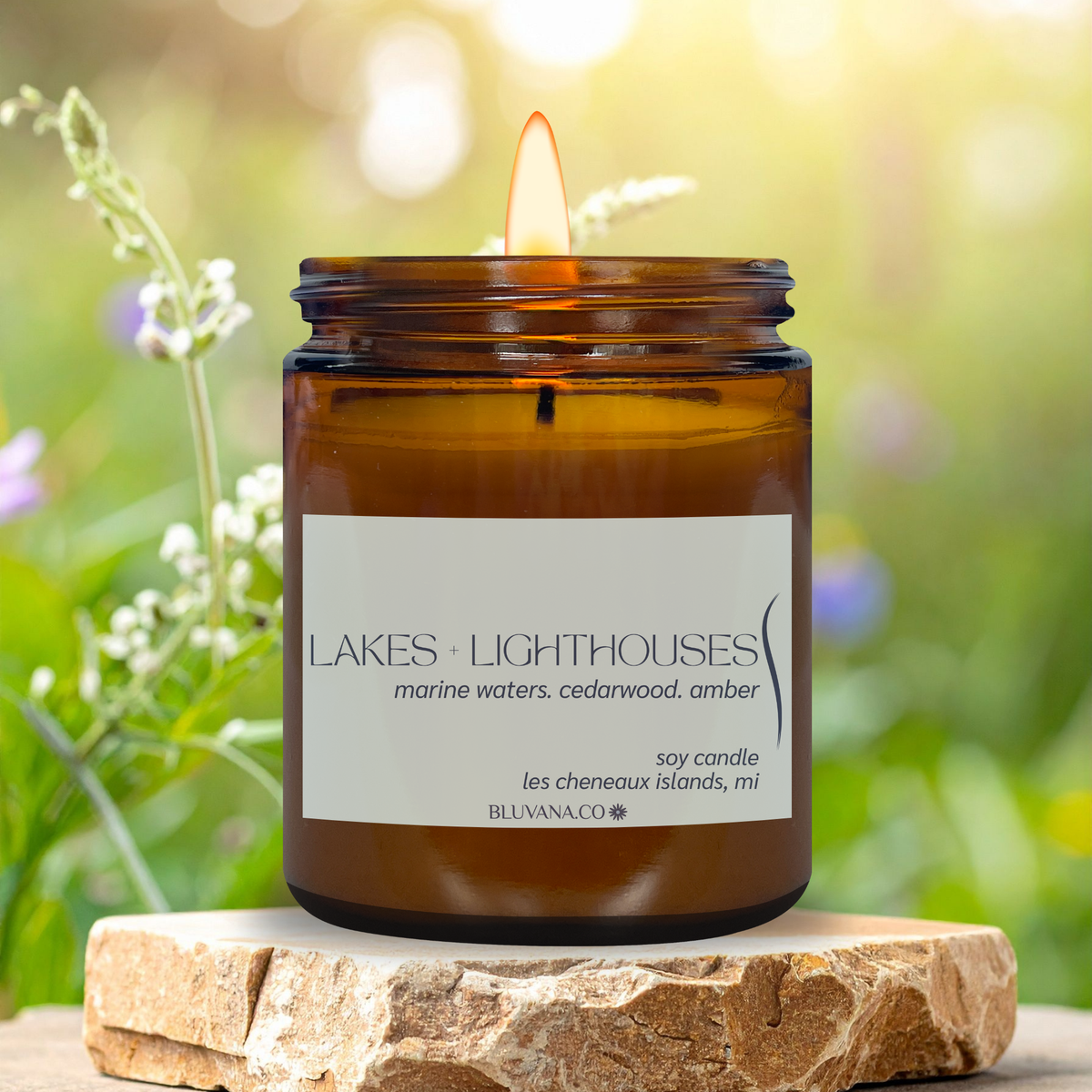Amber jar candle with lakes and lighthouses featuring notes of marine waters, cedarwood, and amber. Situated in a nature inspired setting with florals and rock. 
