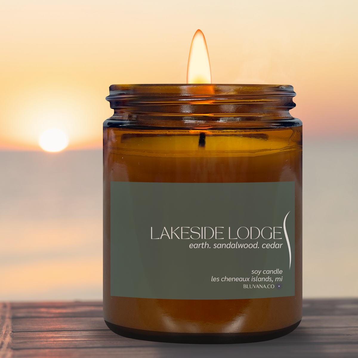 A lit amber glass jar candle with a dark olive green label reading 'LAKESIDE LODGE earth. sandalwood.