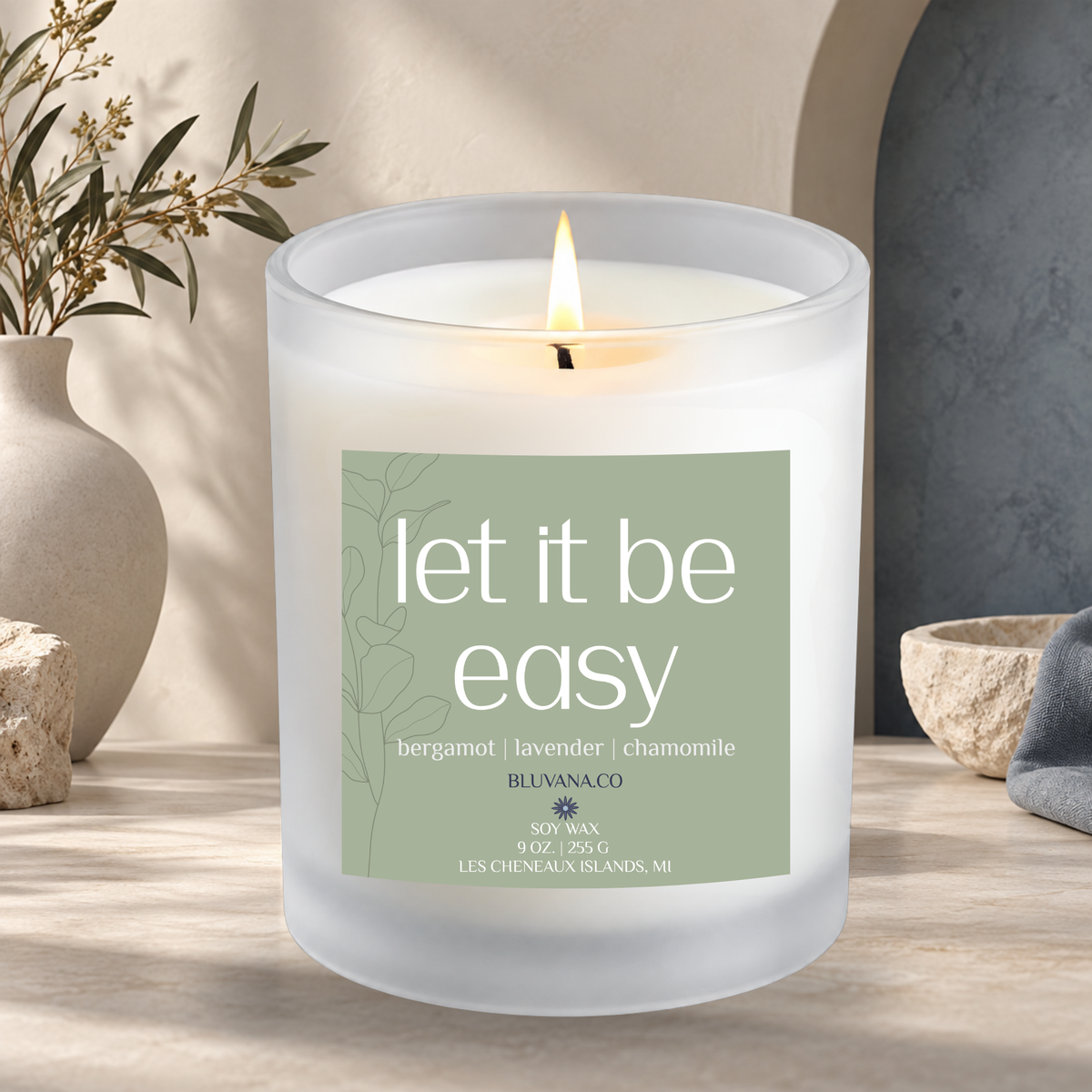 Image of a white frosted glass vessel with flame burning sitting on a modern countertop with ceramic vase in background. Label is a sage green that reads Let It Be Easy with scent notes of bergamot, lavender, and chamomile.