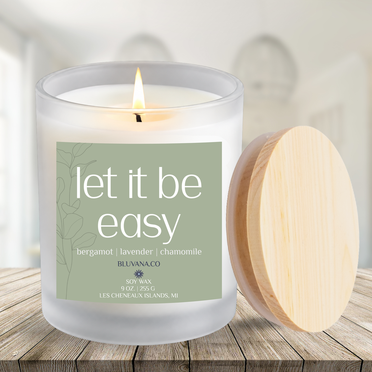 Image of a white frosted glass vessel with flame burning sitting on a modern countertop with ceramic vase in background. Label is a sage green that reads Let It Be Easy with scent notes of bergamot, lavender, and chamomile.