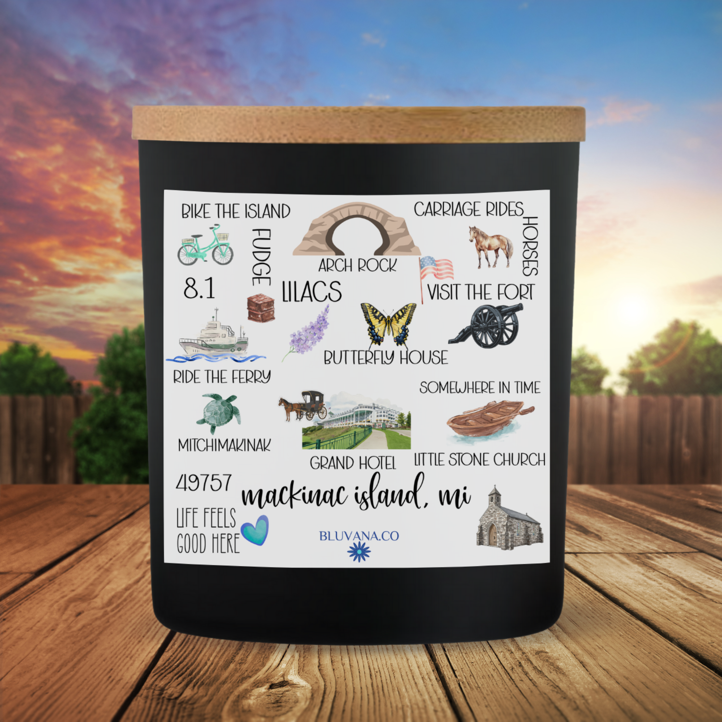 Black candle with illustrations and text about Mackinac Island, MI, on a wooden surface with a scenic background.