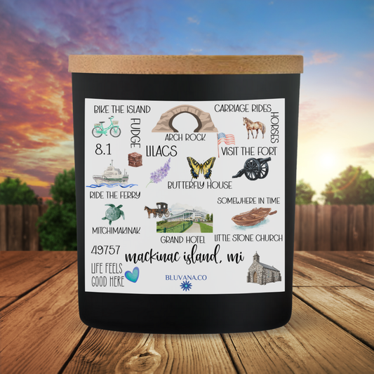 Black candle with illustrations and text about Mackinac Island, MI, on a wooden surface with a scenic background.