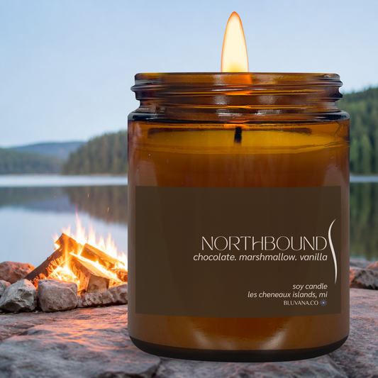 Candle in an amber jar with a label 'Northbound' by a campfire by a lake.