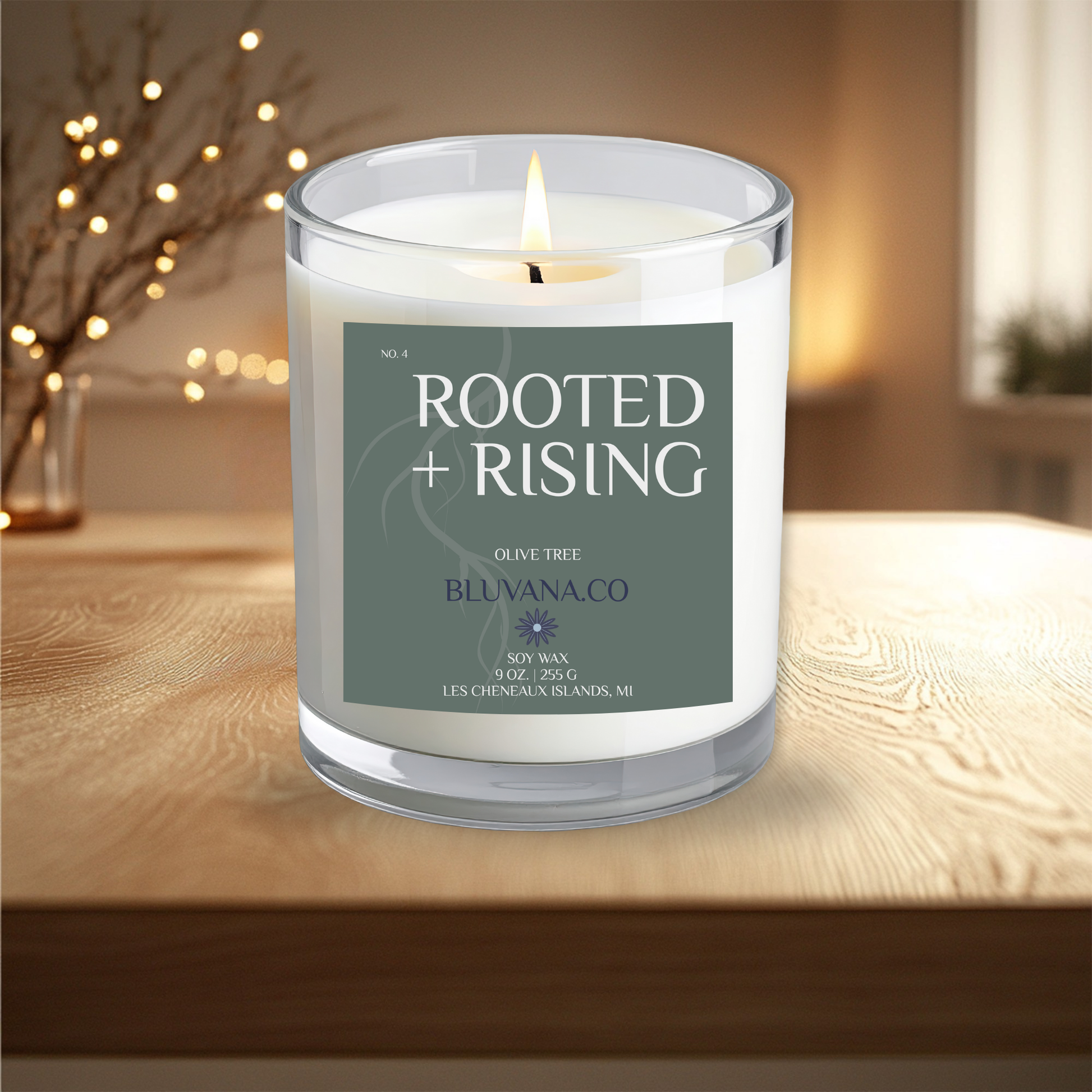 Candle in a glass jar labeled 'Rooted + Rising' on a wooden surface with a blurred background