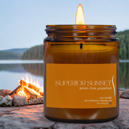 Candle labeled 'Superior Sunset' by a campfire with a scenic background