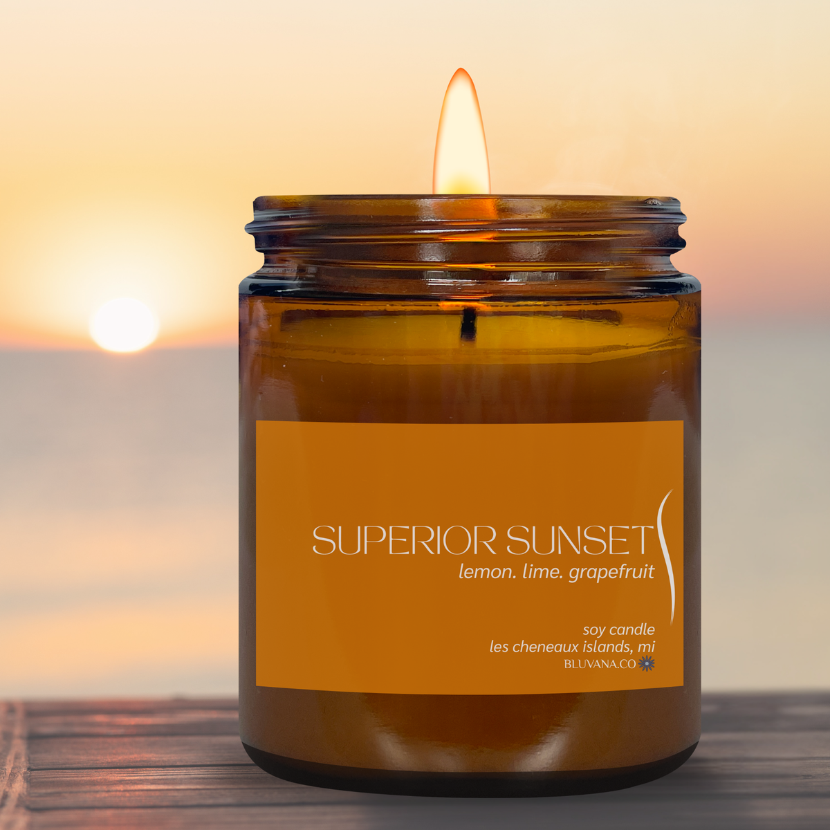 Amber jar candle with orange label that reads Superior Sunset. Sitting on dock with sunset in background. 
