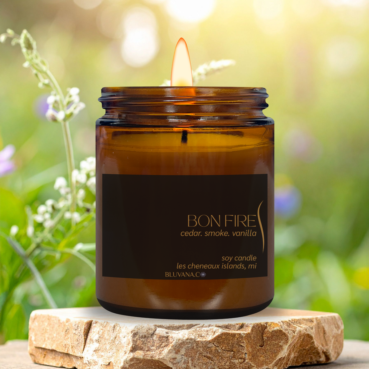 An amber glass jar candle with a lit wick sits on a rustic stone slab outdoors. The label reads 'BON FIRE cedar, smoke, vanilla' and 'BLUVANAC'.